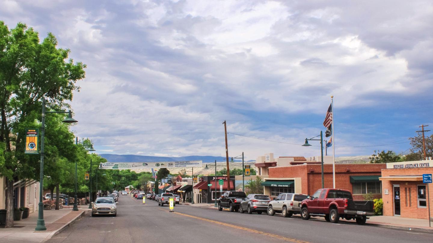 Small town street with parked cars, cloudy sky, and trees lining the road.