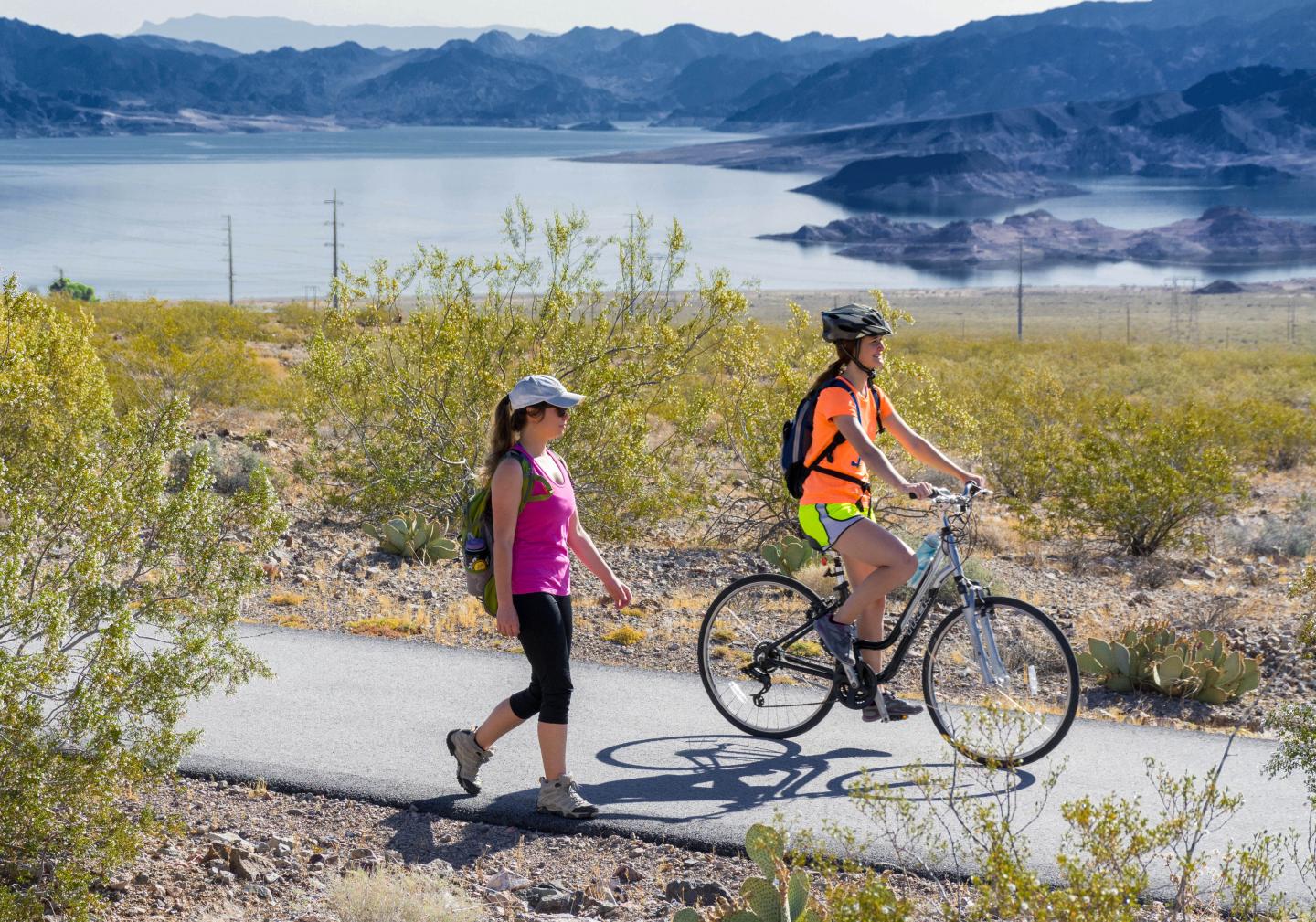 Cyclist and walker on sunny path by a lake with mountains in the background.
