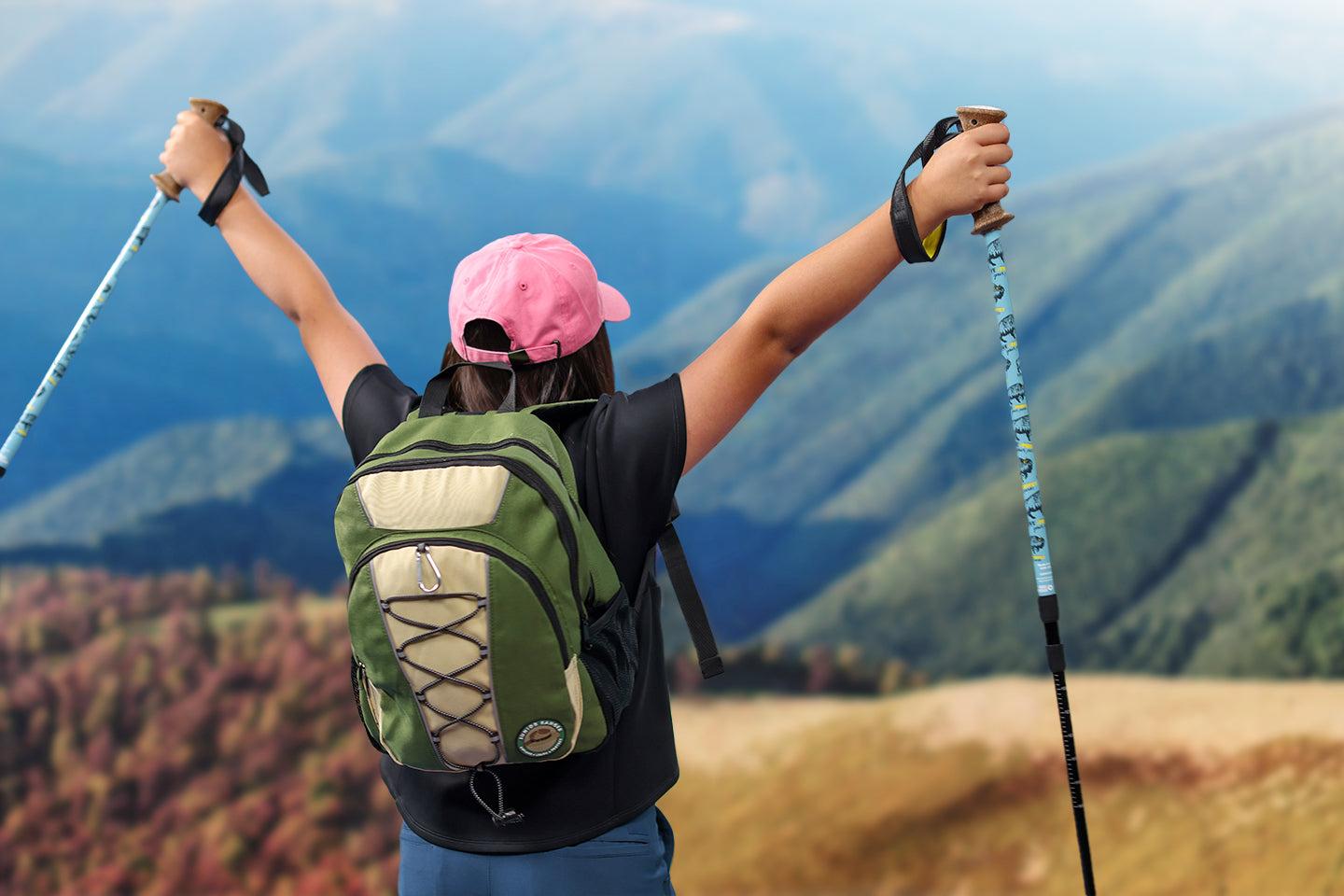 Hiker with pink hat raises arms atop mountain, holding trekking poles.
