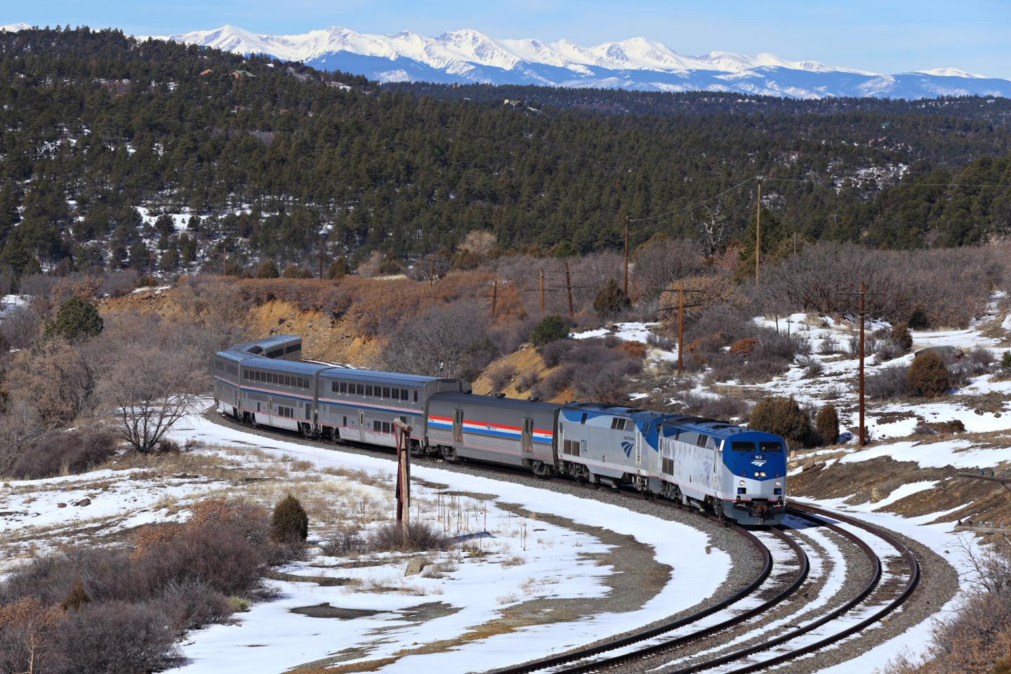 Train on snowy tracks curving through a forested landscape with mountains in the background.