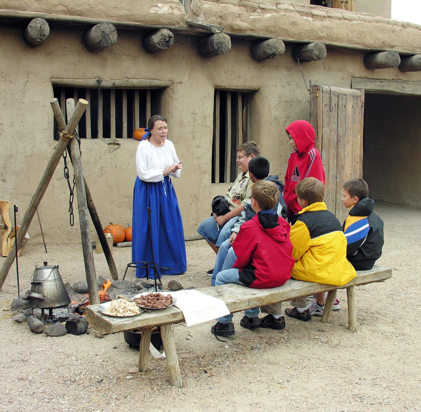 A woman in historical attire talks to attentive children by a fire pit.