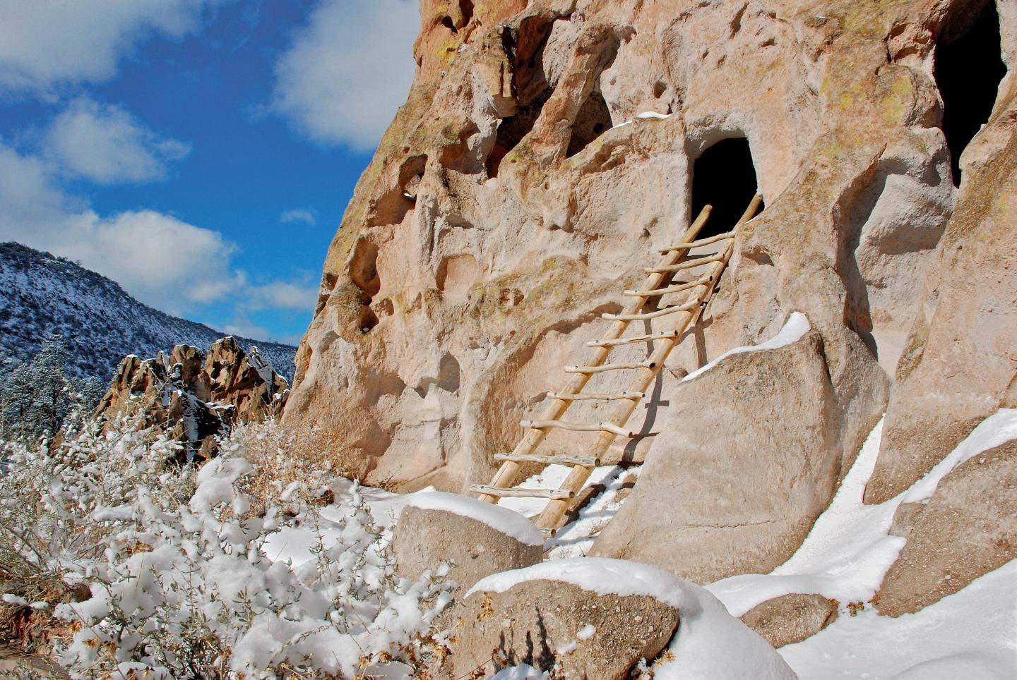 Ladder leading to cave entrance in rocky cliff, snowy landscape under a blue sky.