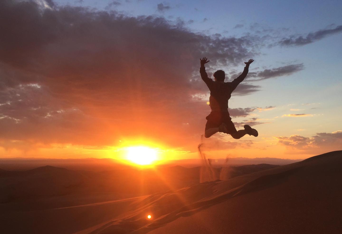 Silhouette of person jumping on a sand dune at sunset.