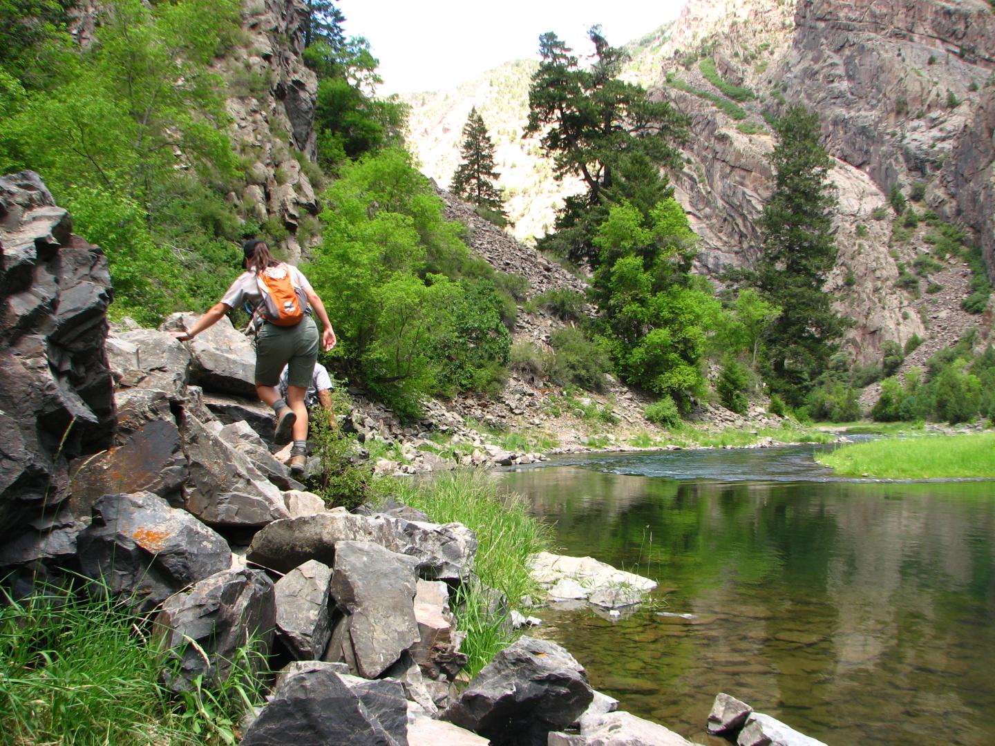 Hiker climbing rocks beside a calm river in a canyon.