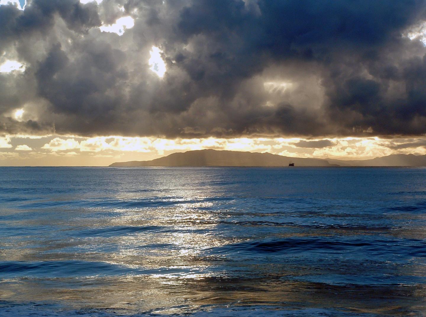 Sunset over ocean with dark clouds, distant land, and glistening water.