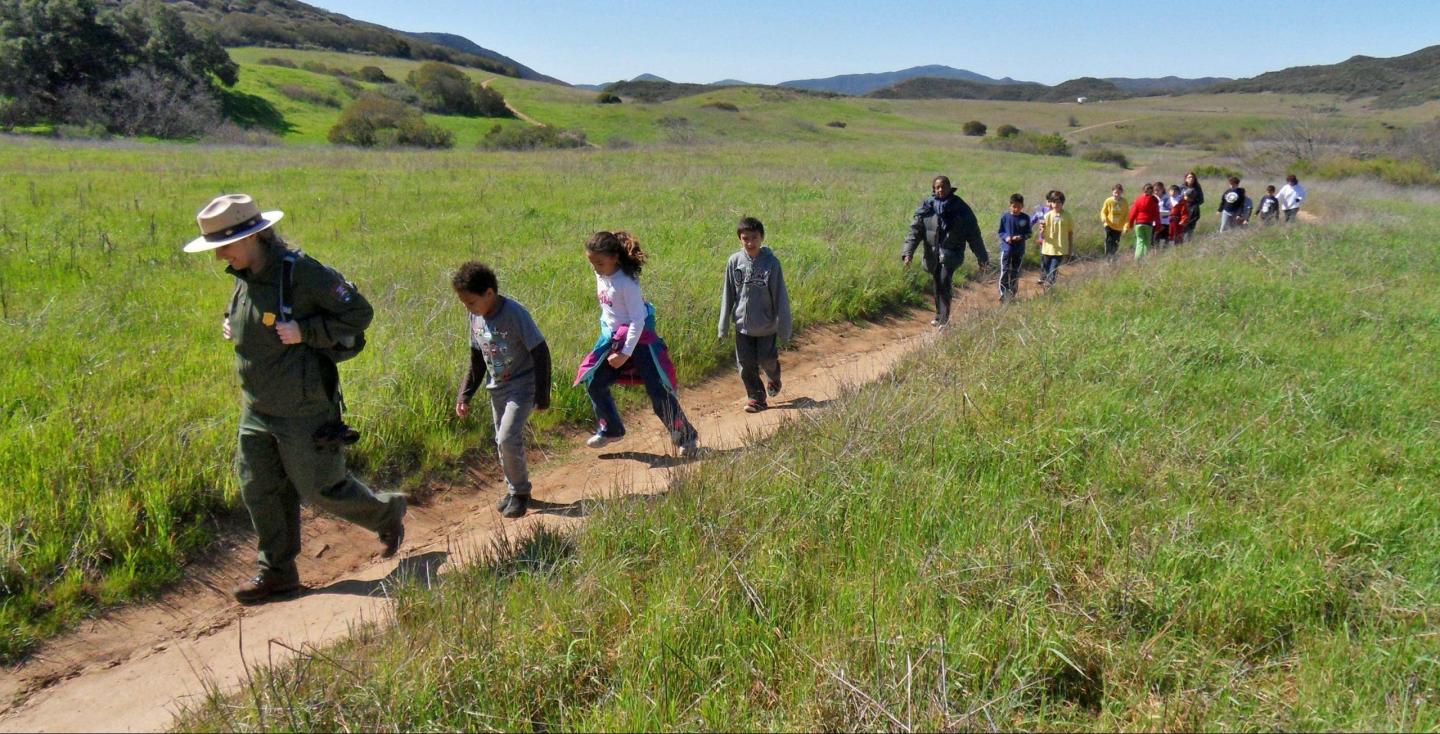 Children hiking on a trail led by a park ranger in a grassy field.