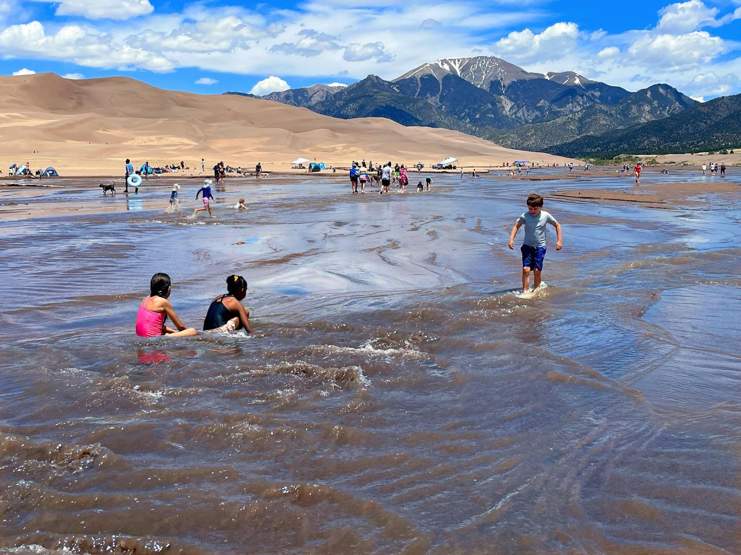 Children play in shallow water at a dune with mountains in the background.