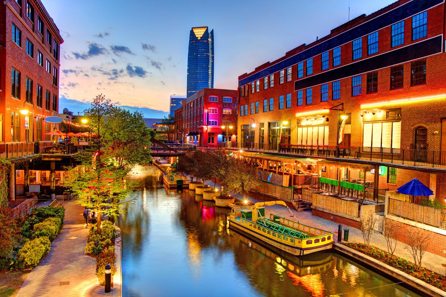 Colorful Oklahoma City riverside with lit buildings at dusk, reflecting in the water.