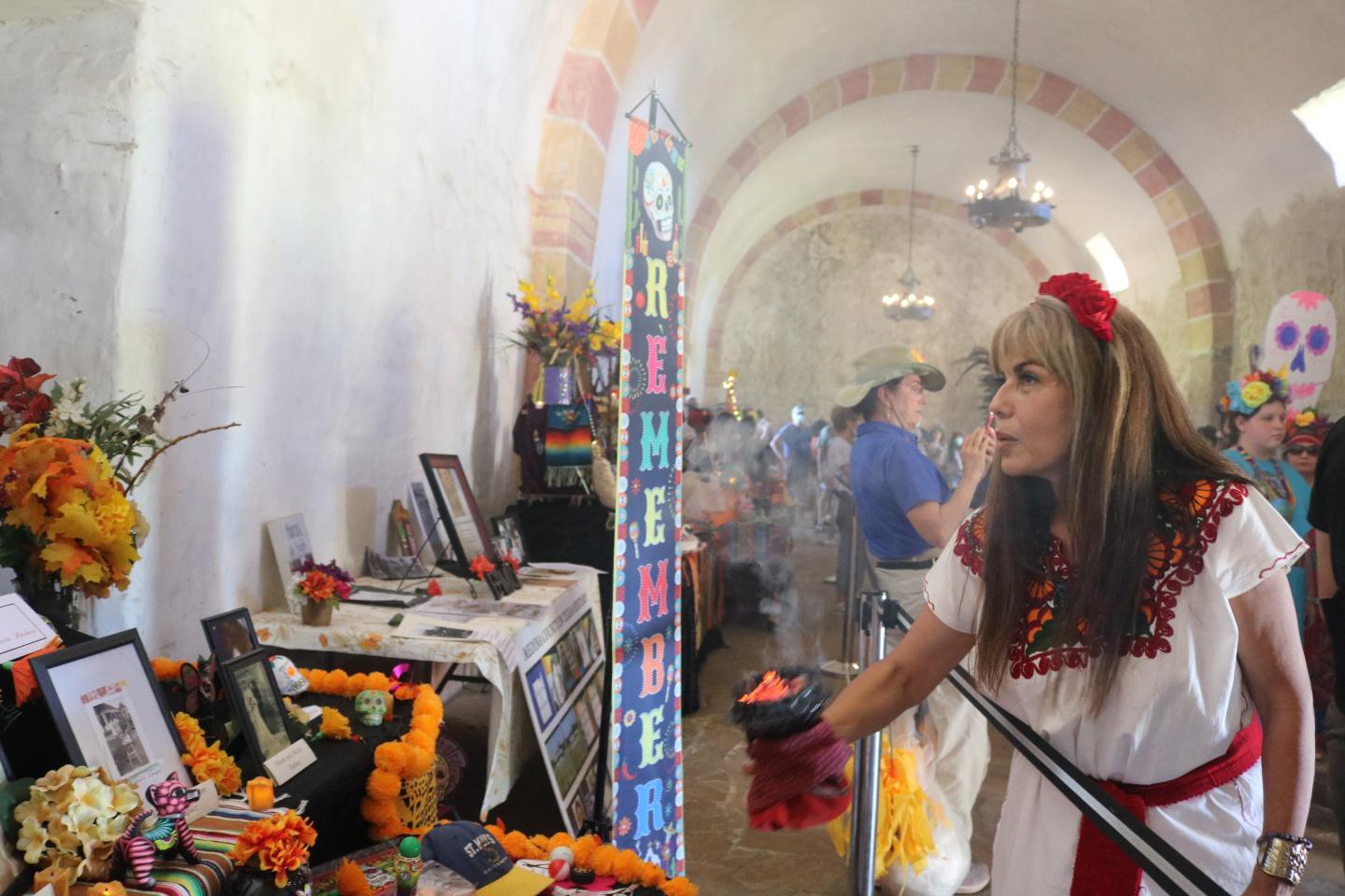 Woman in traditional dress at a Fiesta San Antonio altar with flowers and candles.