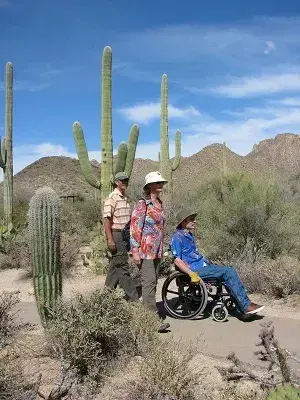Three people on a desert trail with cacti; one uses a wheelchair.