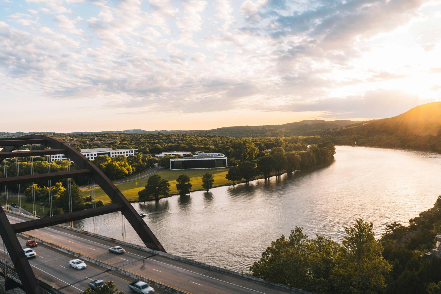 River by a bridge at sunset with cars, trees, and a partly cloudy sky.