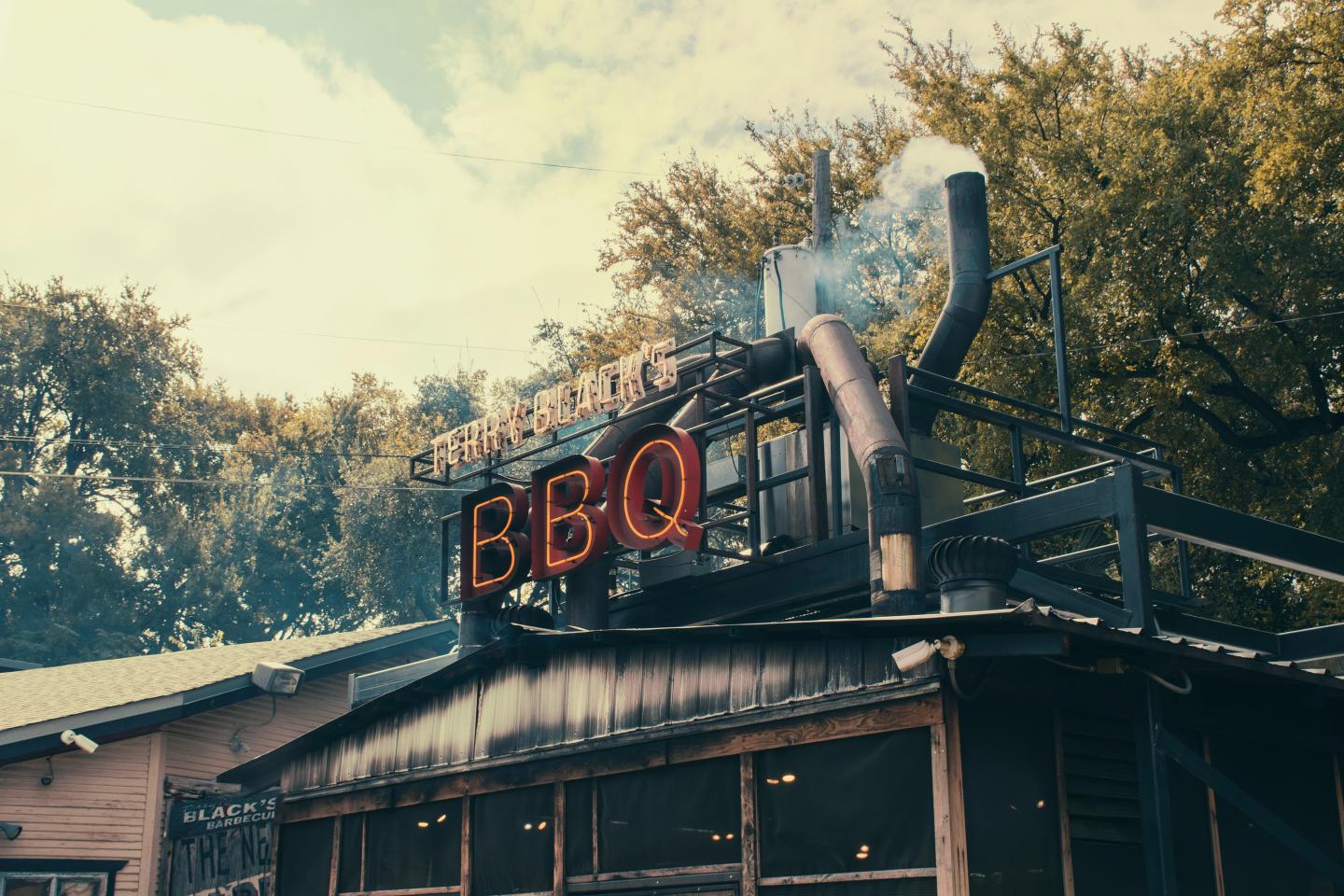Barbecue restaurant with neon BBQ sign and smoke rising near trees.