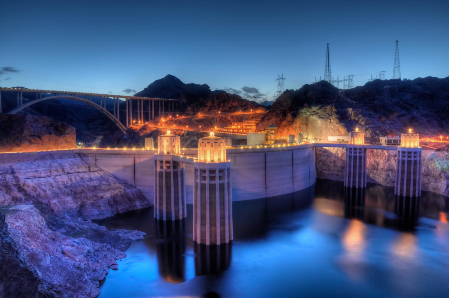 Hoover Dam at sunset, illuminated with lights and a blue sky backdrop.