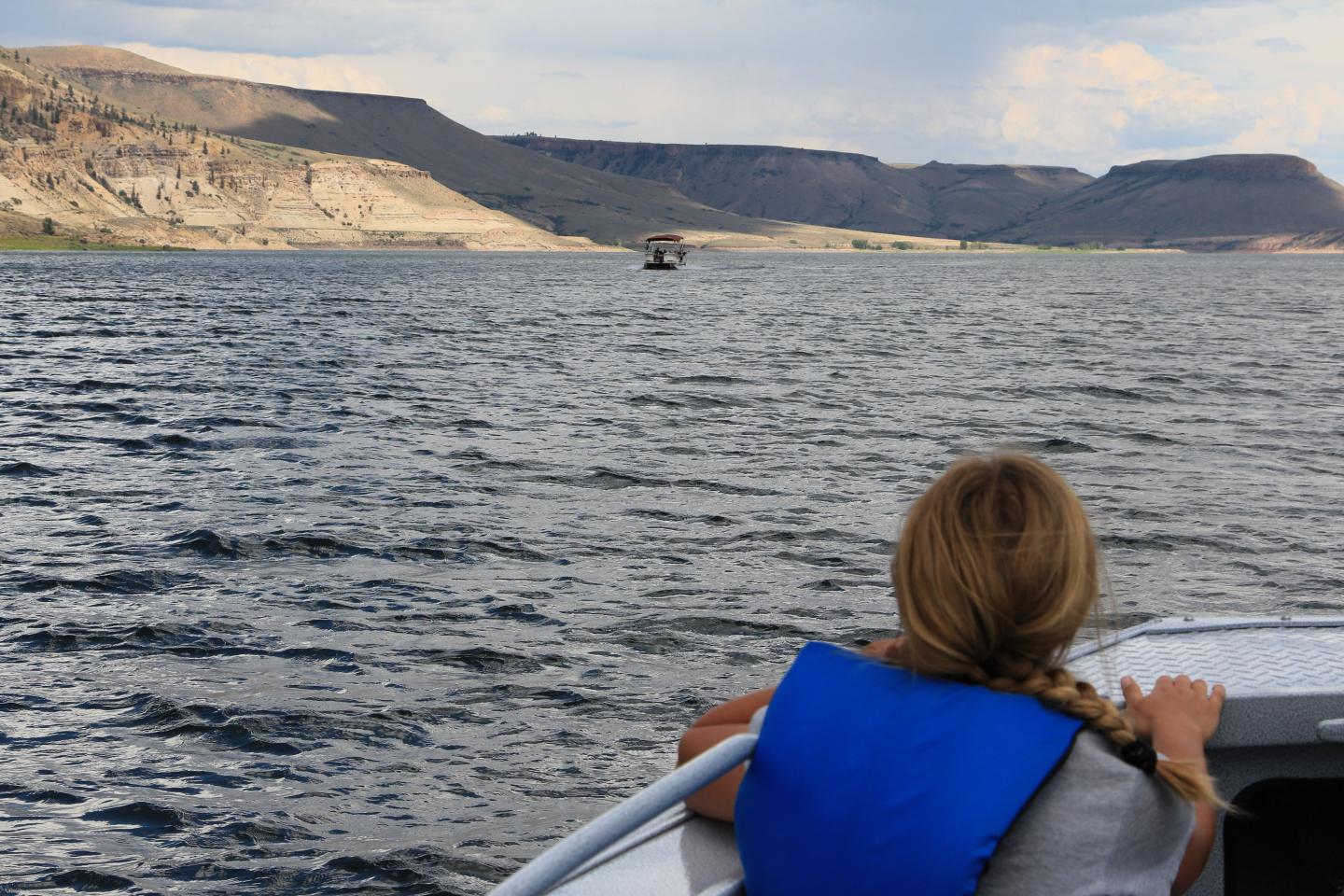 Girl with a braid in a blue life jacket on a boat, looking at distant cliffs on a lake.