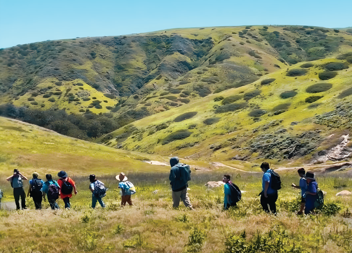 Hikers walking on a grassy trail with green hills in the background.