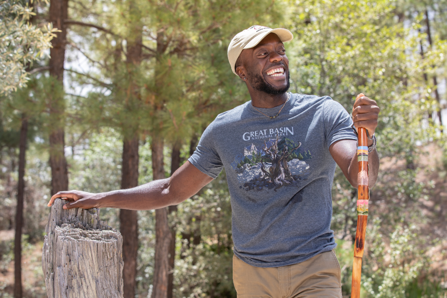 Man smiling, hiking in a forest with a walking stick, wearing a cap and graphic T-shirt.