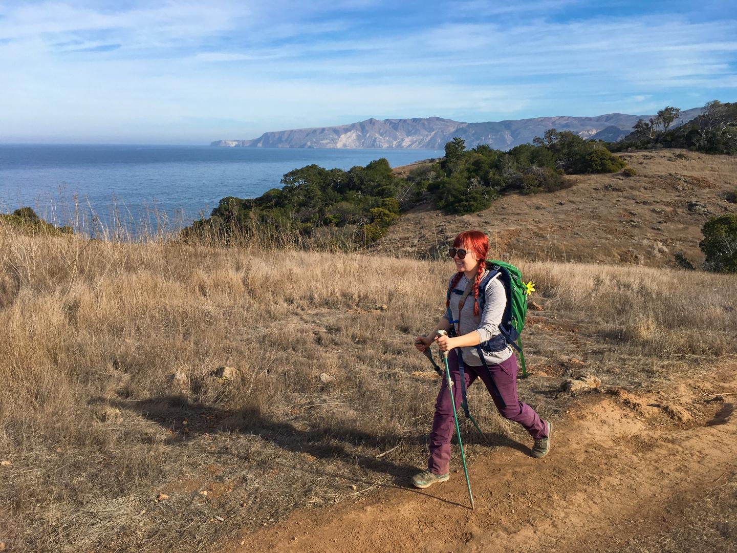 Hiker with a backpack walking along a coastal trail on a sunny day.