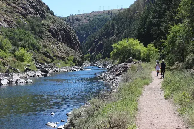 Trail along a scenic river with people walking and green hills around.