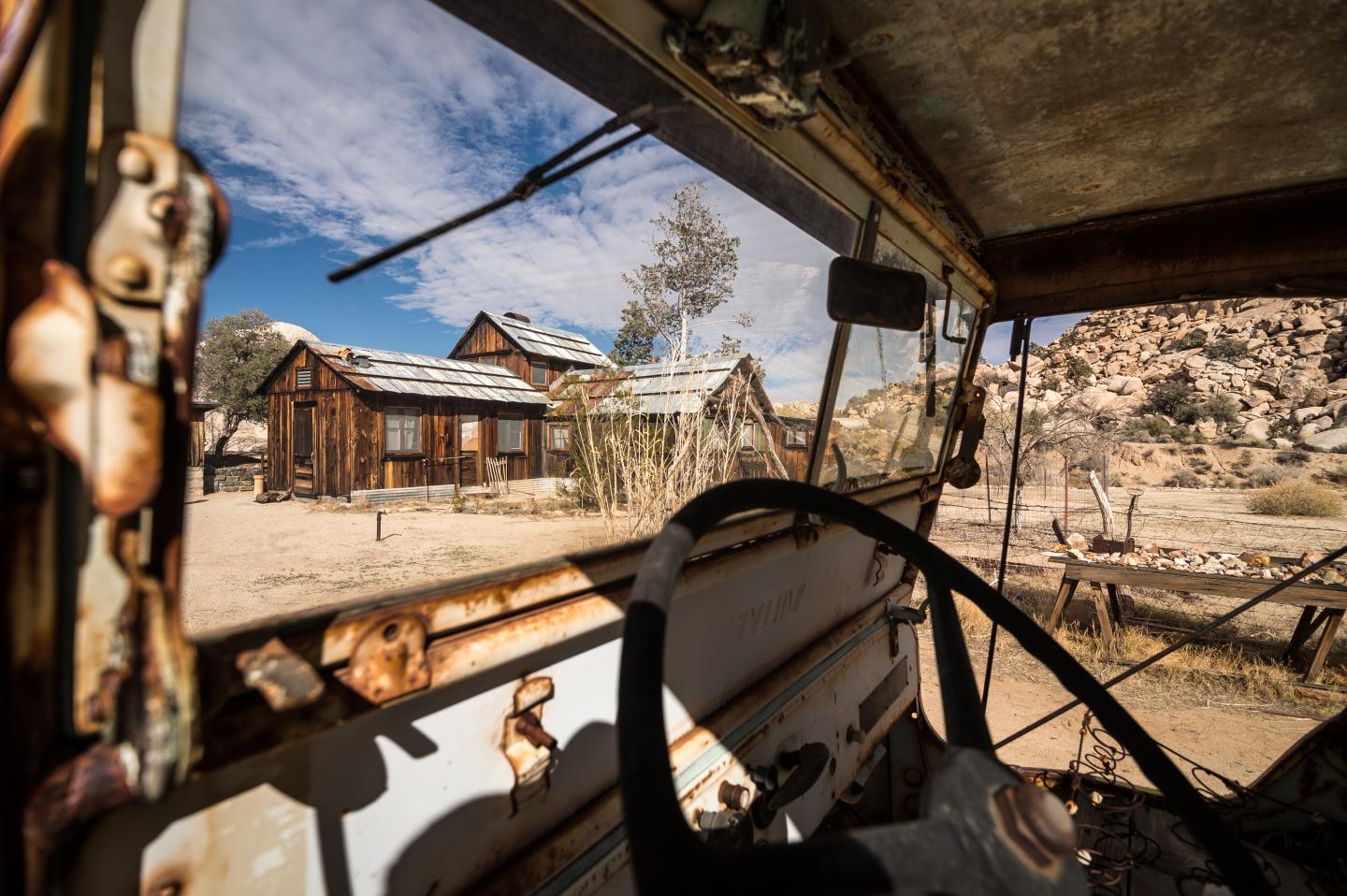 Rusty car interior overlooking old wooden cabins in desert landscape.