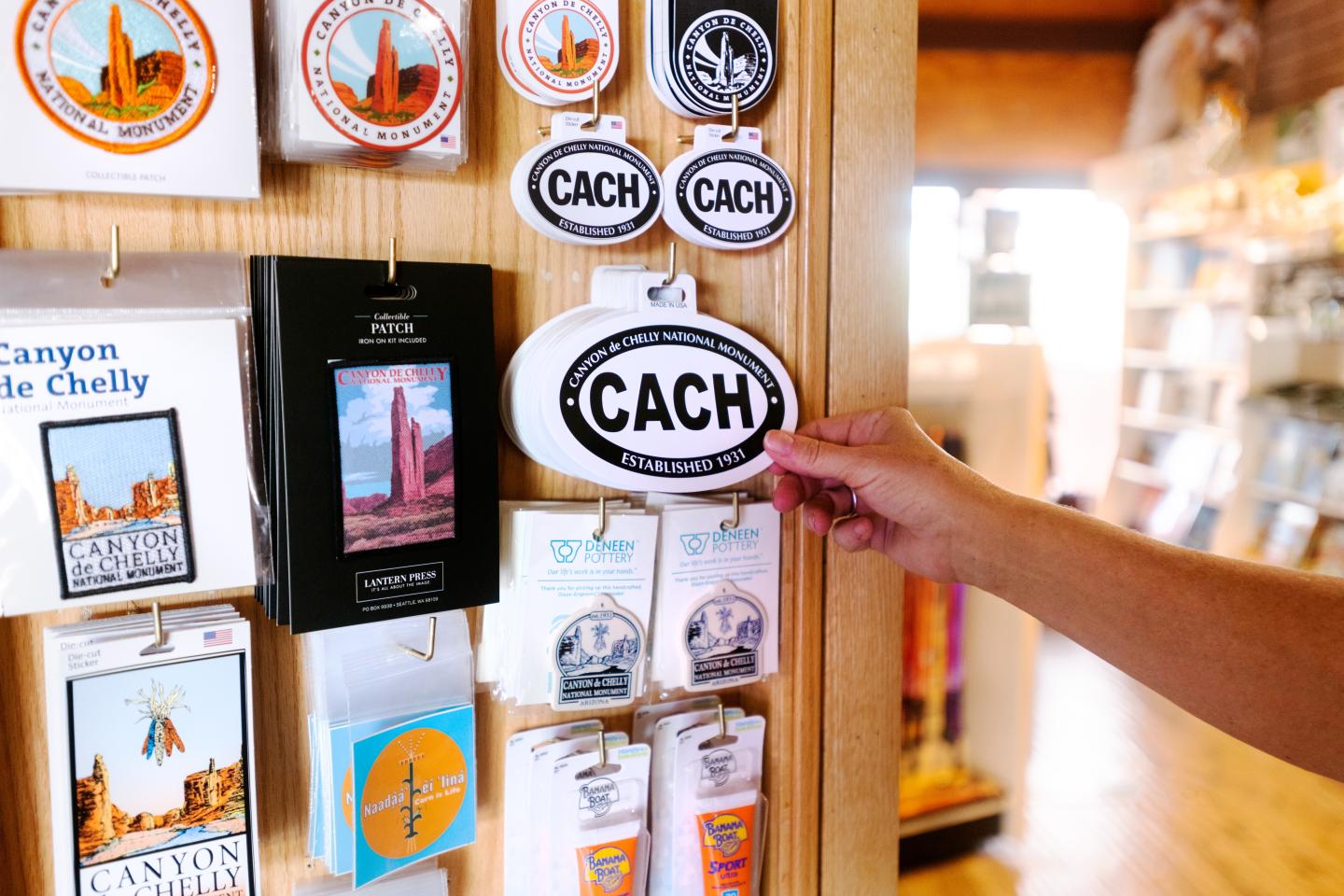 Hand holding a CACH sticker near a display of postcards and stickers in a store.