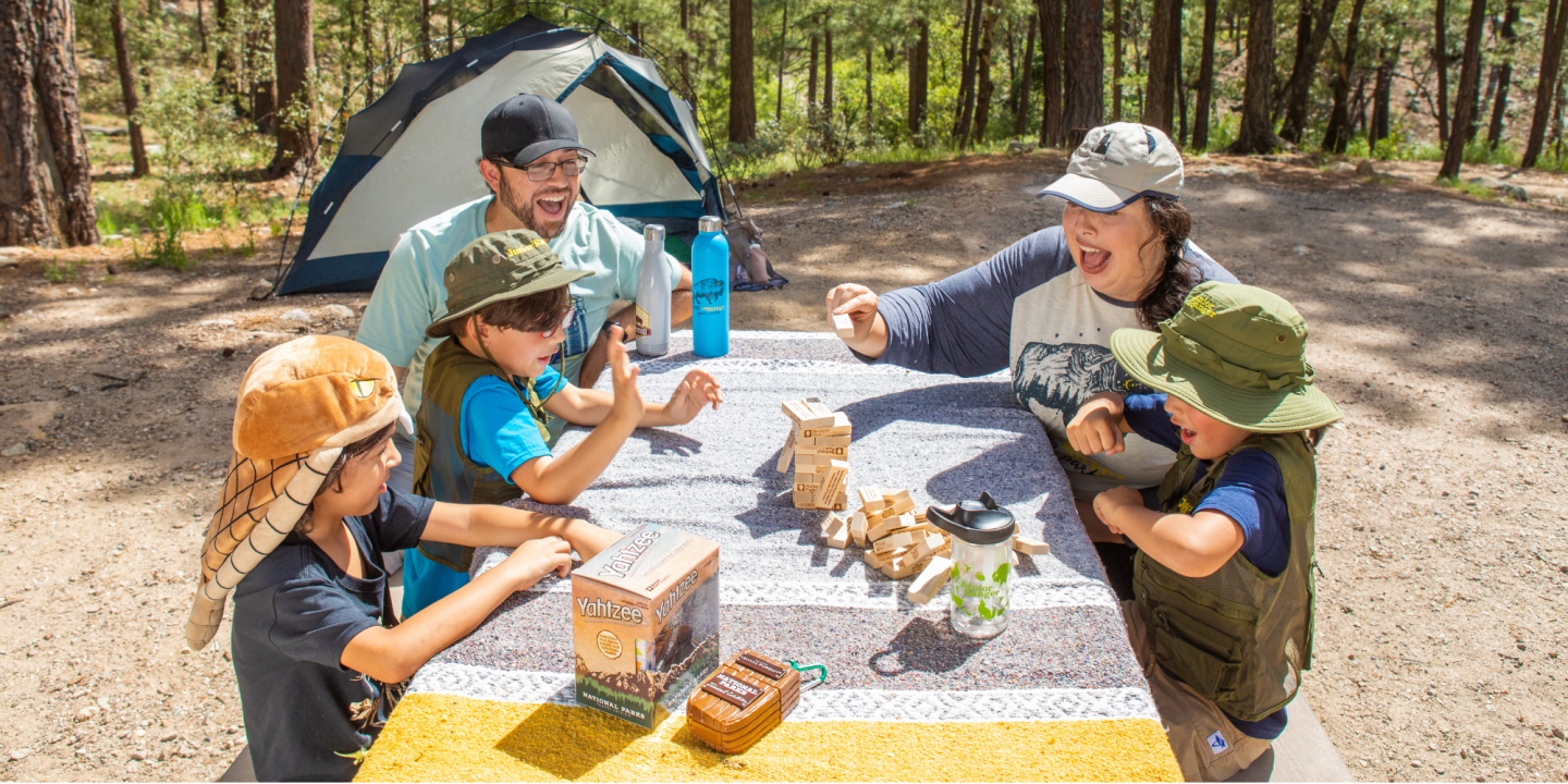 Family playing a game at a picnic table near a tent in the woods.