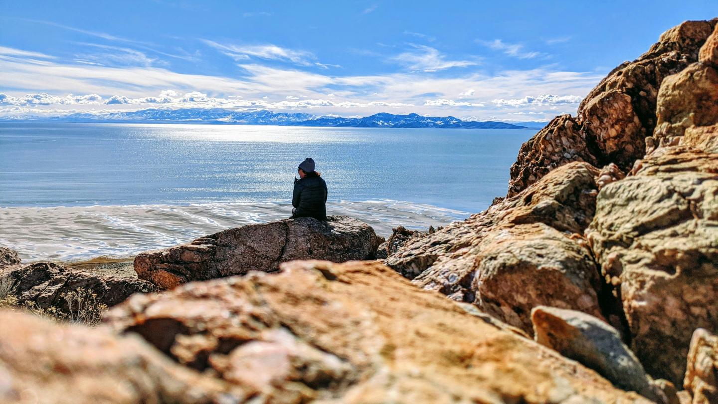 Person sitting on rocky shore, looking at the ocean under a blue sky.