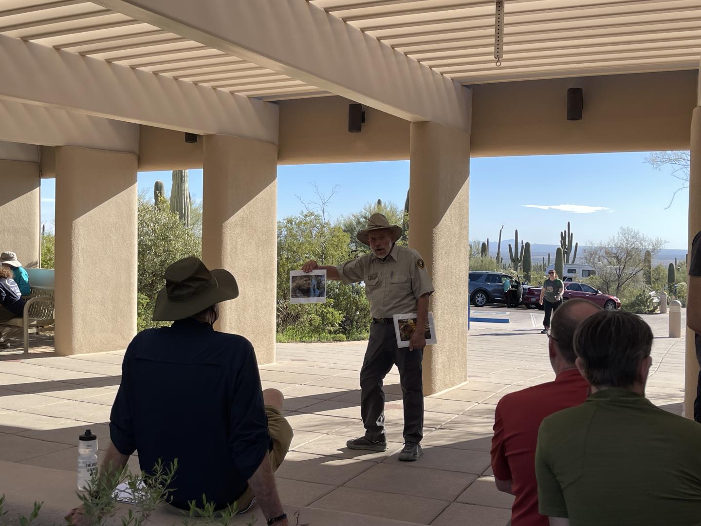 Man in hat lecturing to seated group outdoors under pergola.