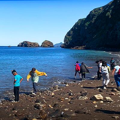 People walking along a rocky beach with blue sea and cliffs.