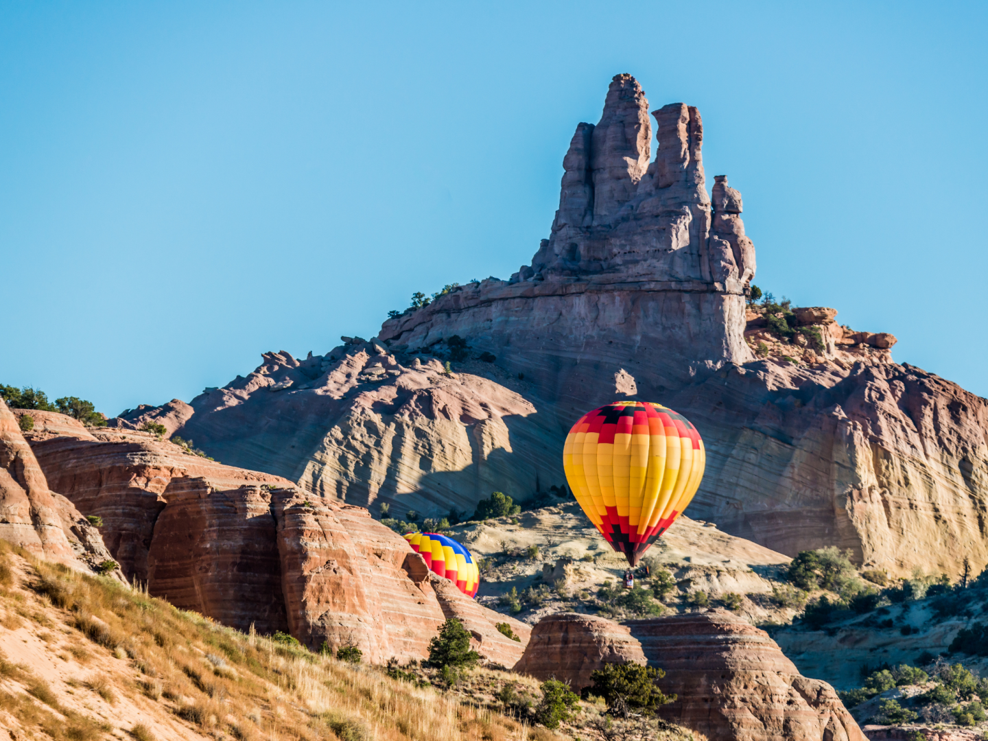 Hot air balloons float near rocky desert cliffs under a clear blue sky.