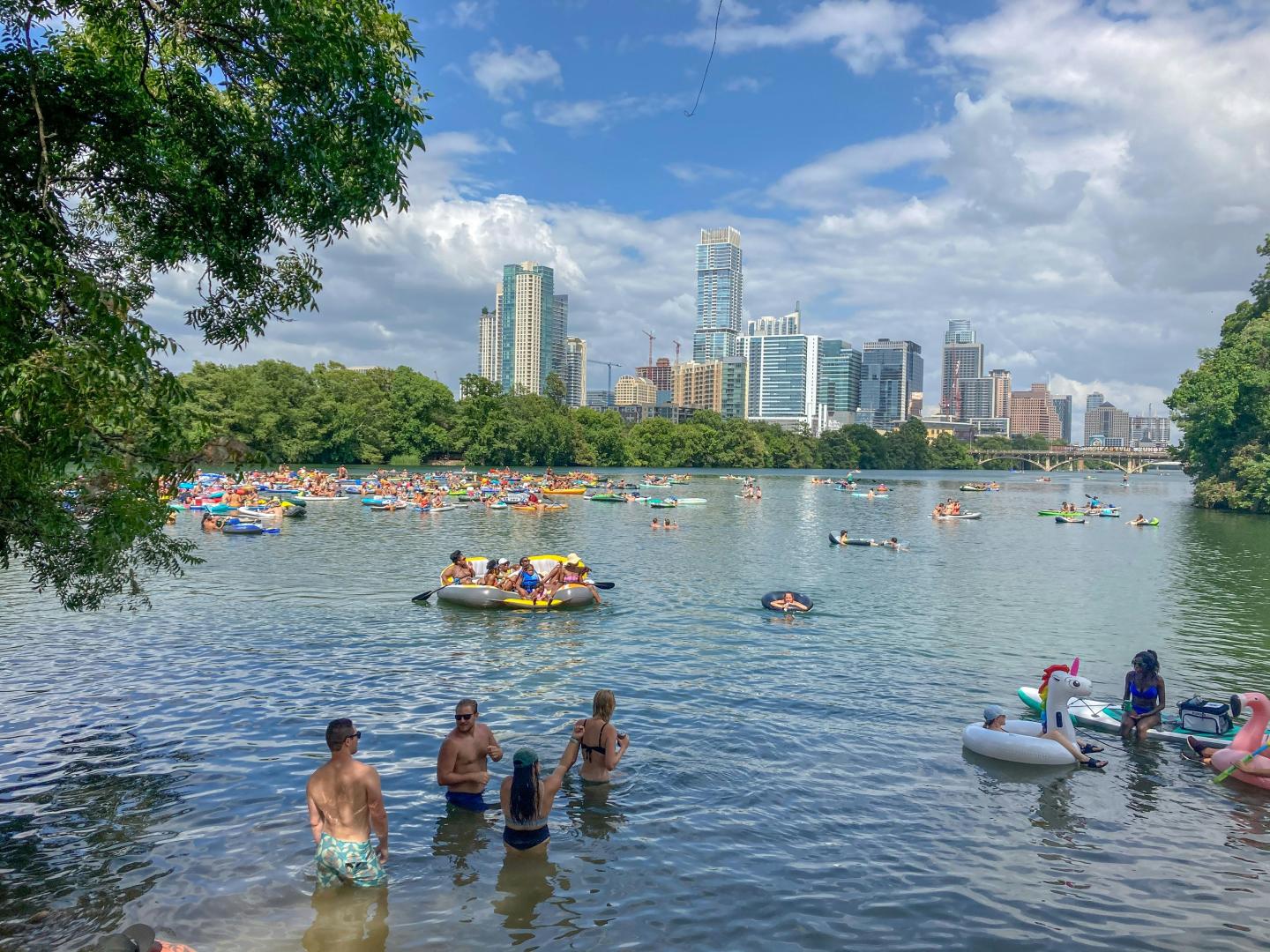 People in a lake with floats, city skyline in the background, under a partly cloudy sky.