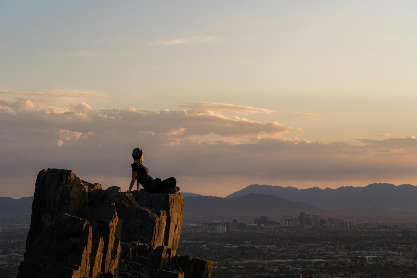 Person sitting on a rock at sunset, city and mountains in the background.