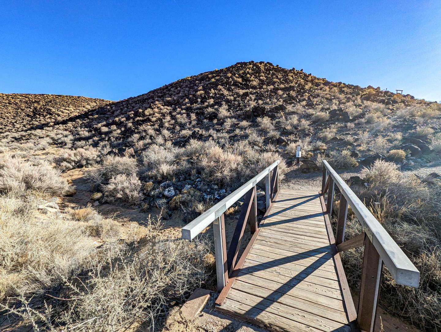 Wooden footbridge leading to a dry, bushy hillside under a clear blue sky.