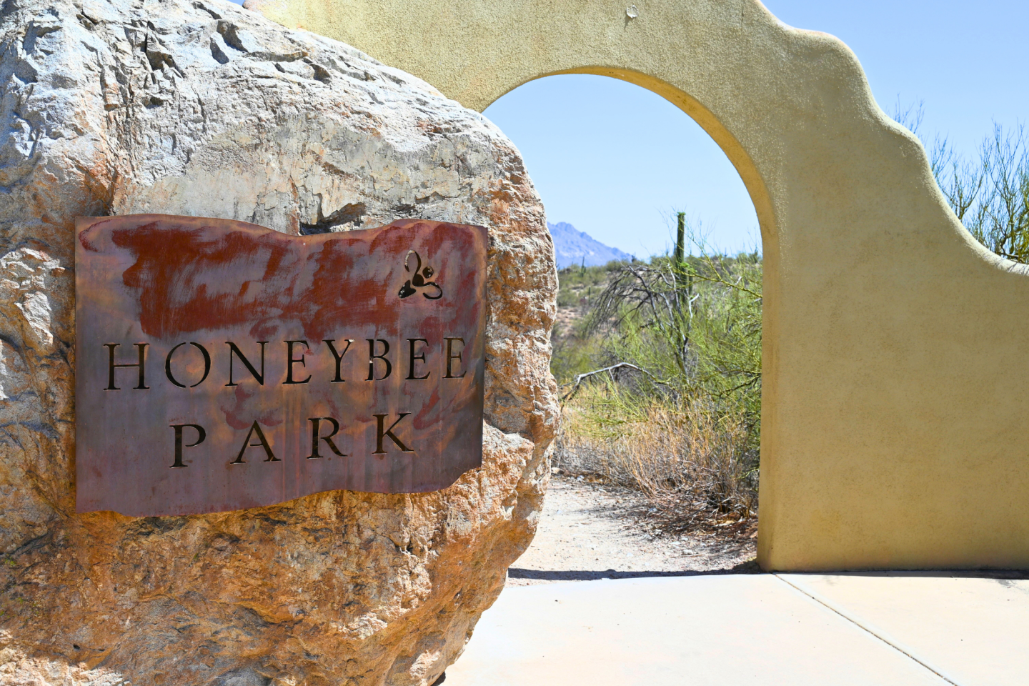 Honeybee Park sign on a rock, desert plants, archway, mountain in background.
