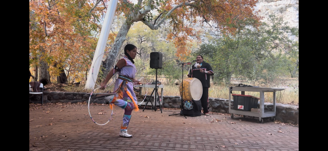 Young hoop dancer outdoors near a drummer, surrounded by autumn trees.