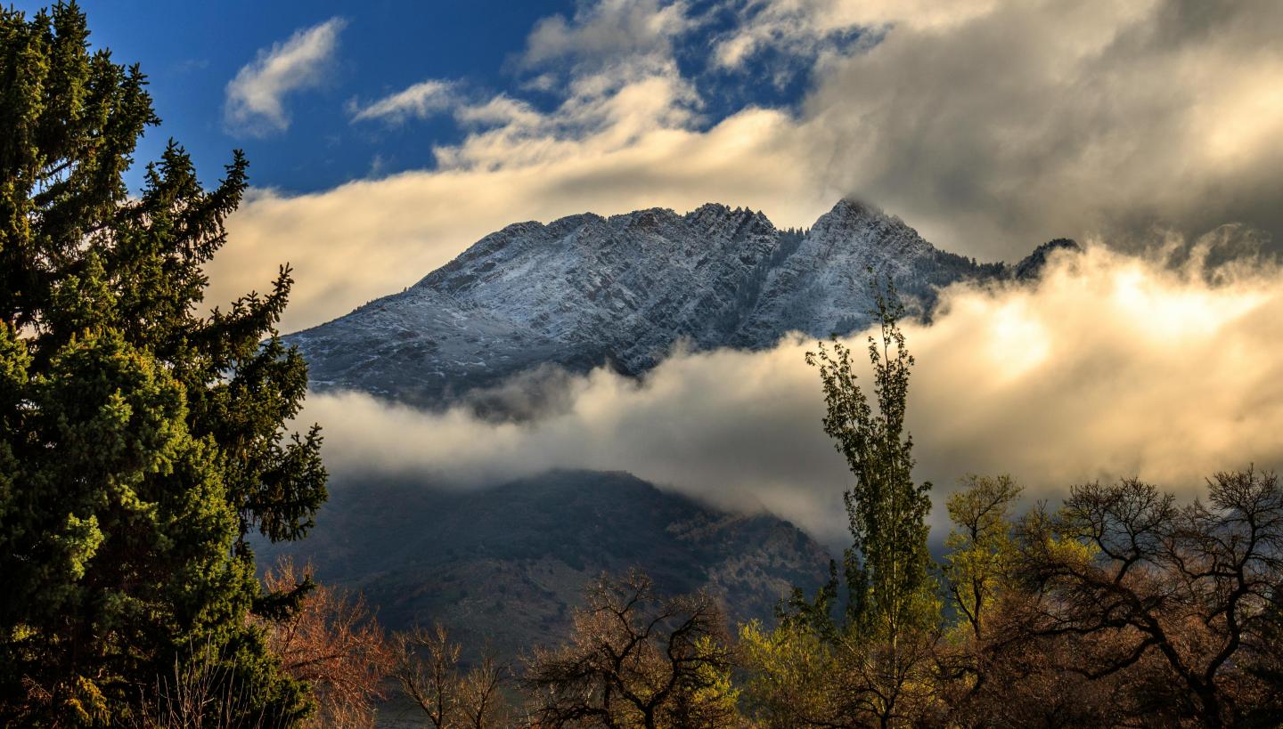 Snowy mountain peak under blue sky, surrounded by clouds and trees.
