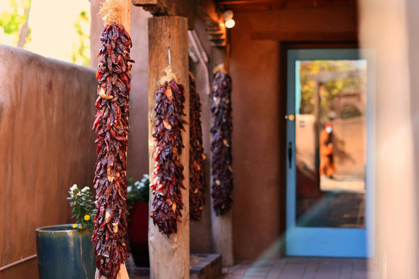 Dried chili ristras hang in a sunlit adobe hallway near a bright blue door.