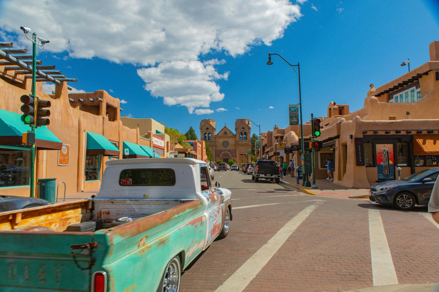 Old truck driving through a sunny, adobe-style street with blue sky overhead.