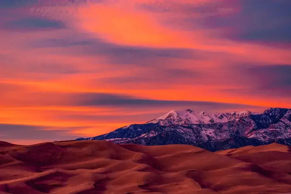 Vibrant sunset over snow-capped mountains and desert sand dunes.