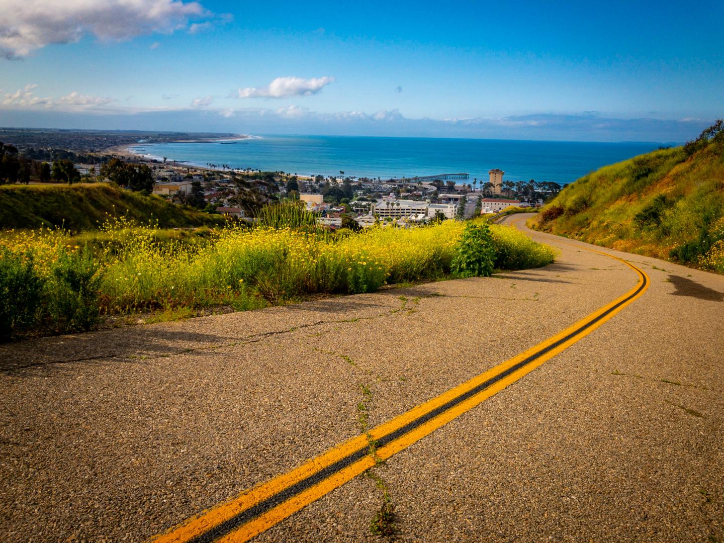 Curving road with city view, lush hills, and blue ocean under a clear sky.