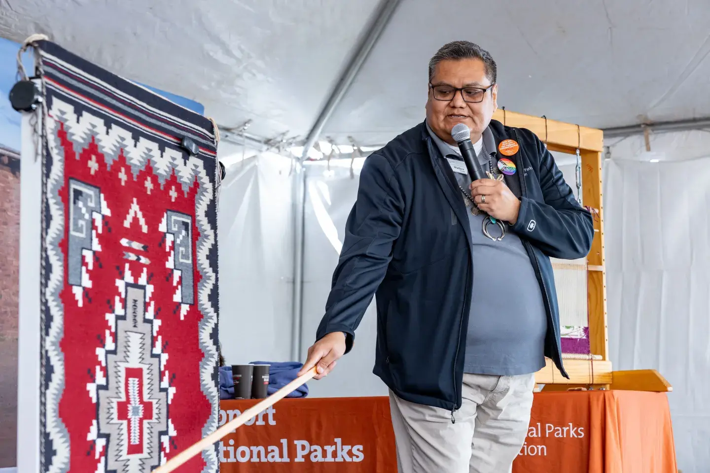 Wallace James Jr speaking into a microphone, pointing at a colorful woven Navajo rug under a tent.