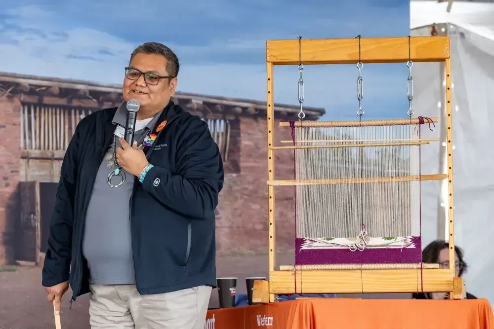 Wallace James Jr speaking next to a loom on a stage at Tucson Festival of Books, outdoor setting.