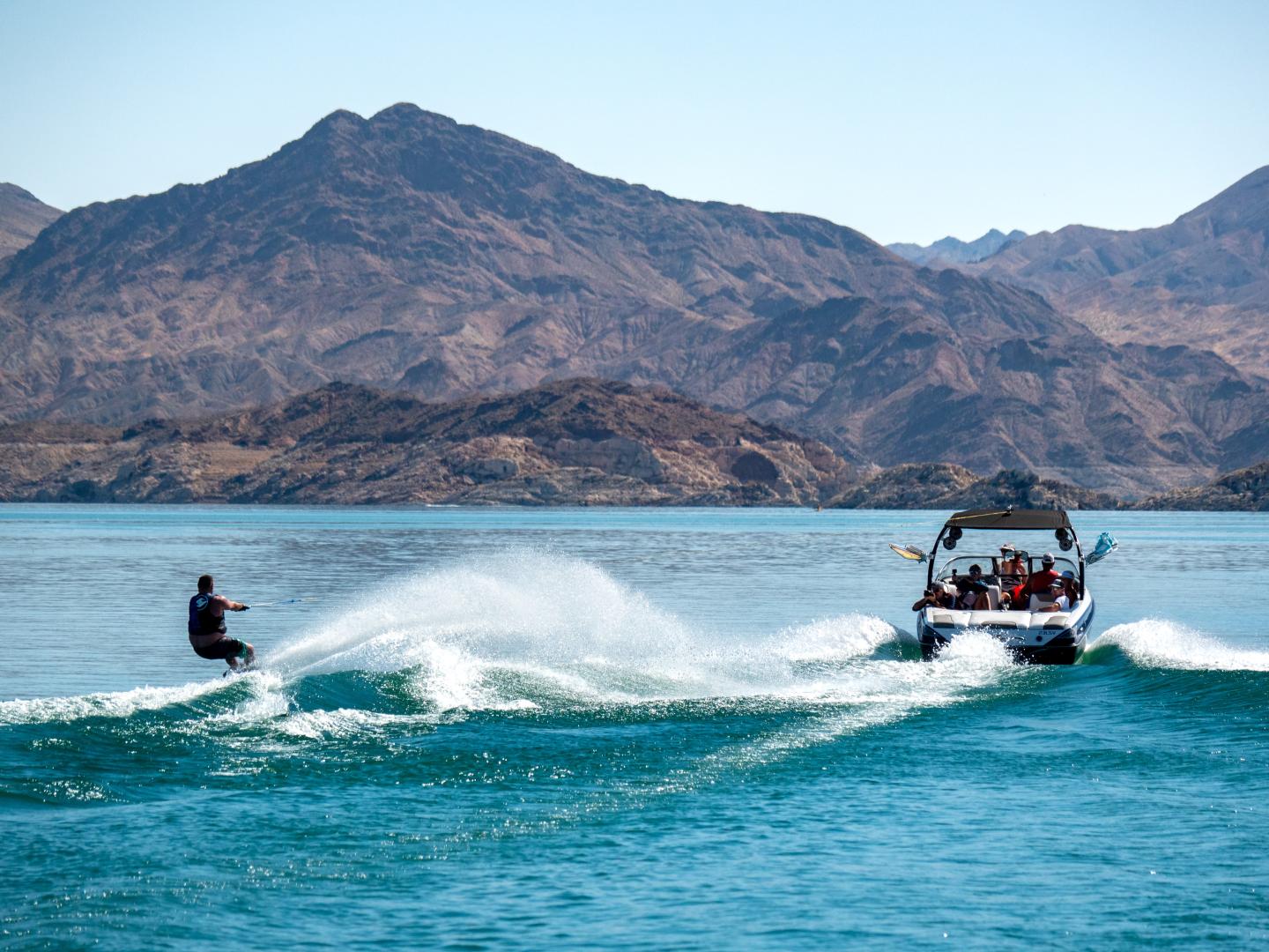 Wakeboarder riding waves behind a speedboat on a lake with mountains in the background.