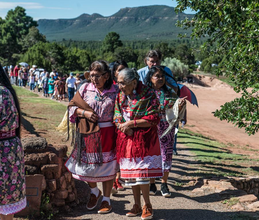 People in colorful traditional attire walking outdoors, mountains in the background.