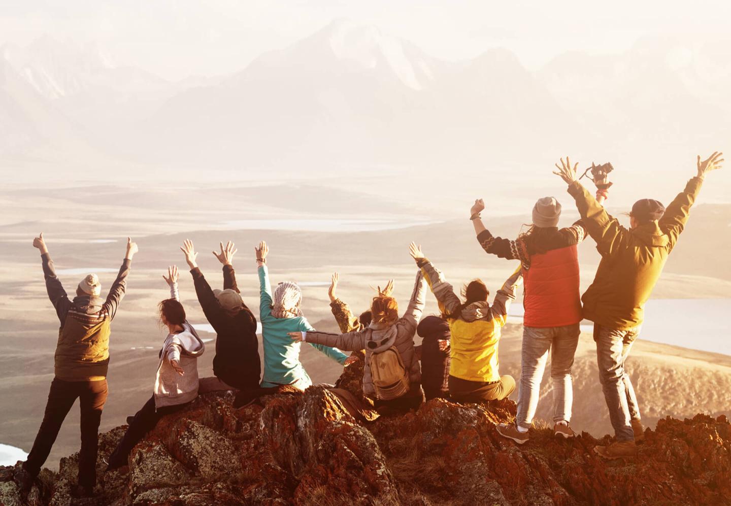 Group of people on a mountain, arms raised, with a scenic background.