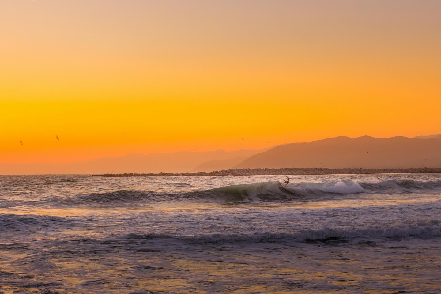 Surfer riding a wave at sunset, with an orange sky and distant mountains.