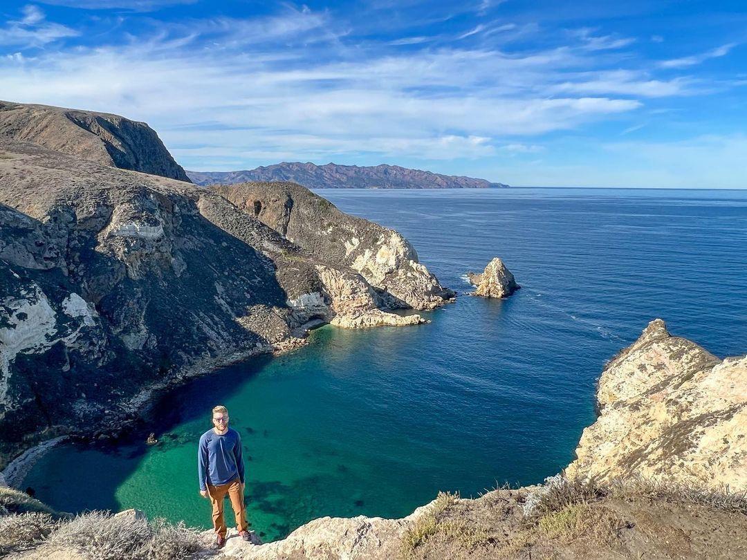 Coastal view with person standing on cliff overlooking a clear blue sea under a bright sky.