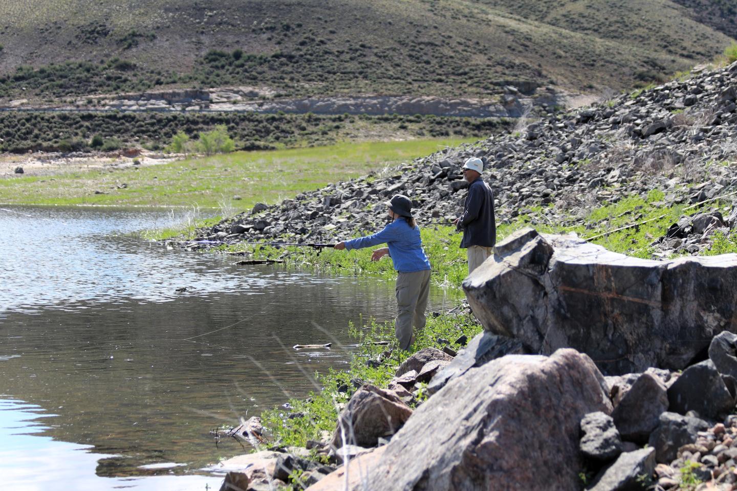 Two people fishing by a rocky lakeside under a clear sky.