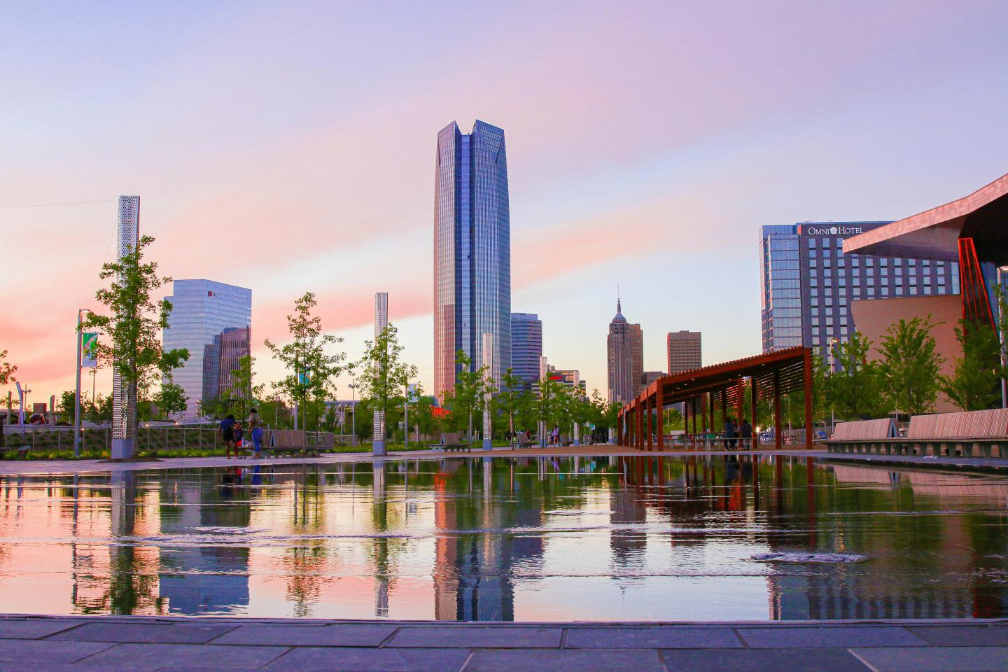 City skyline at sunset with buildings reflected in a calm pond.