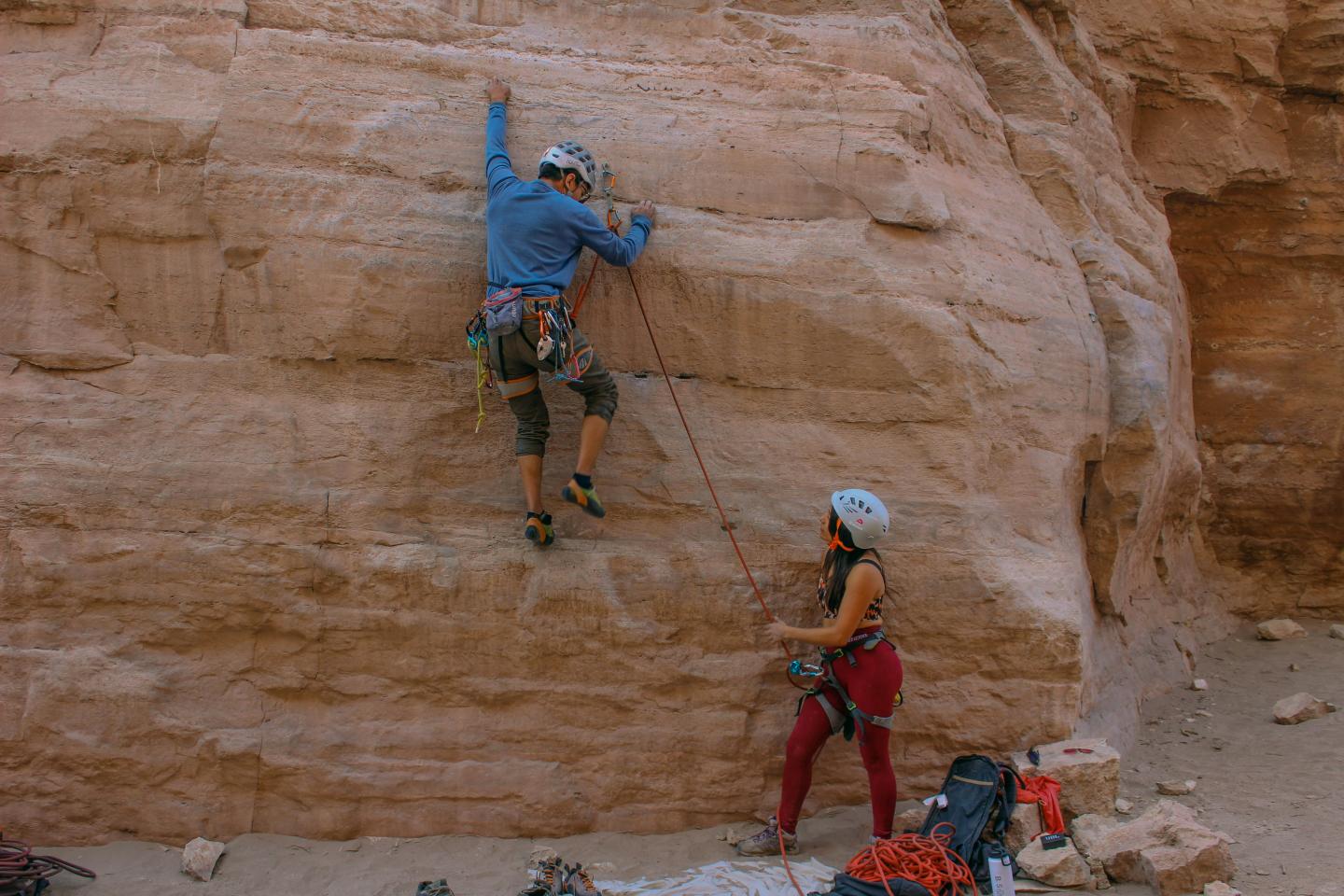 Rock climber scales a sandstone wall, belayer watches, gear nearby.