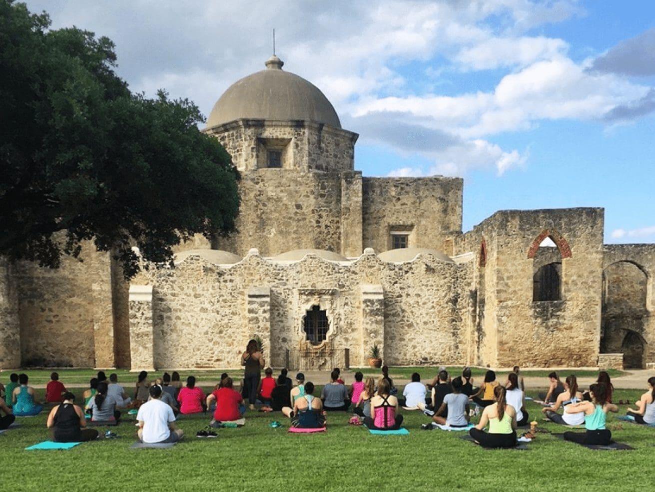 People practicing yoga on grass facing a historic stone building under a blue sky.